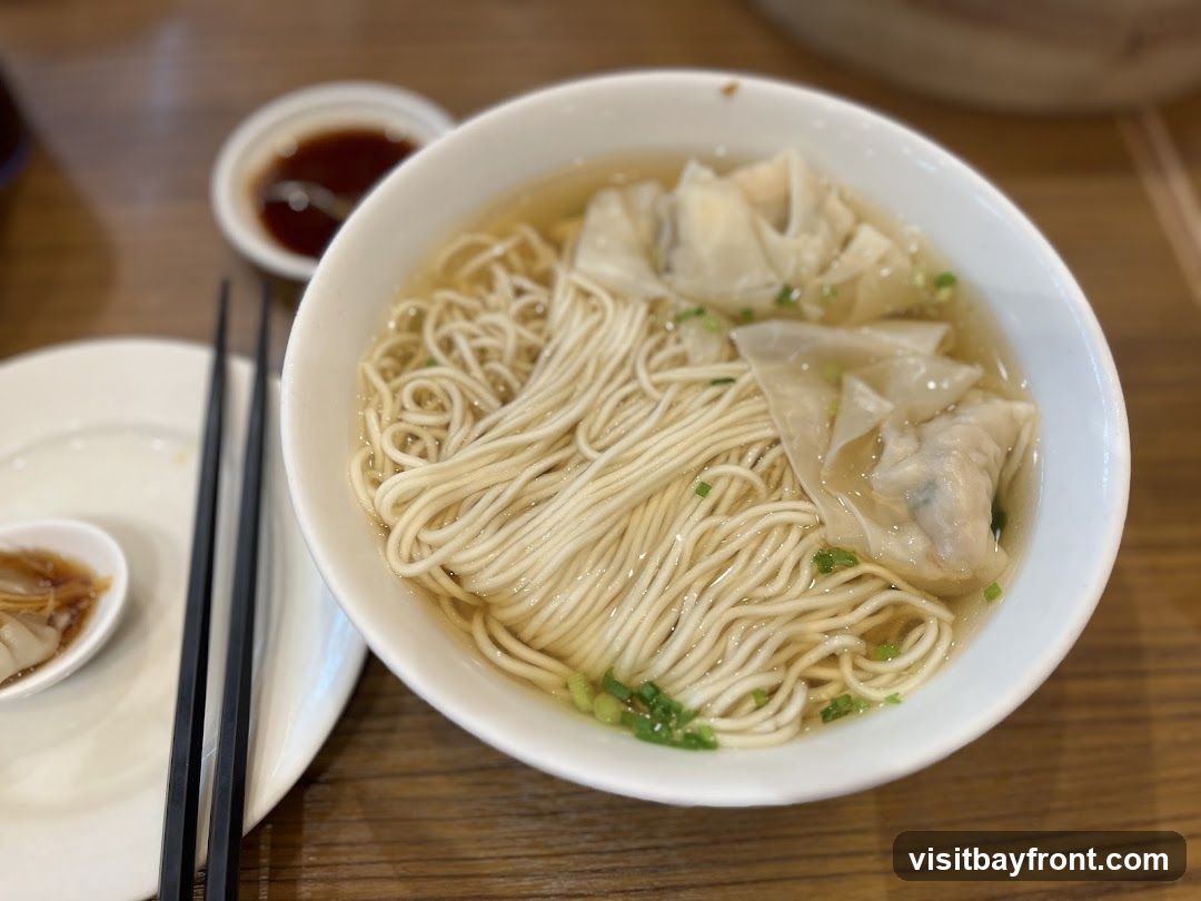 A close-up shot of a bowl of wonton noodle soup. The soup contains thin noodles, several wontons, and chopped green onions. A small dish of soy sauce and a spoon with a wonton are visible on the side.