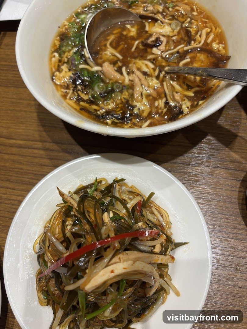 A close-up shot of two dishes of Asian food on a wooden table. In the foreground is a white plate with a cold dish of shredded seaweed, chicken, and noodles, seasoned with chili and herbs. In the background, a white bowl contains hot and sour soup with shredded chicken, mushrooms, and egg. A ladle and spoon are in the soup.