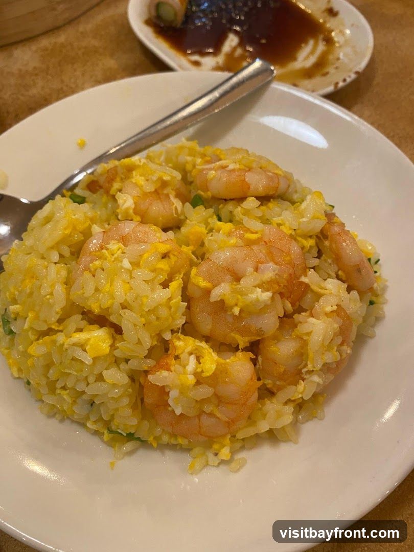 A close-up shot of a plate of shrimp fried rice, with several plump shrimp mixed into the fluffy rice and scrambled eggs. A spoon rests on the side of the plate. In the background, another plate holds a dish with soy sauce and a rolled item.