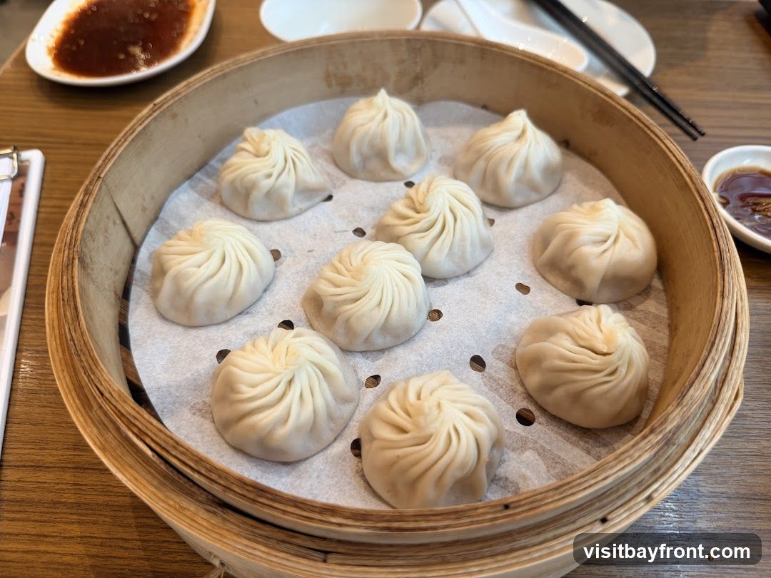 A bamboo steamer basket filled with soup dumplings sits on a wooden table. There are ten dumplings arranged in the basket, each with a pleated top. A small dish of dipping sauce is visible to the right of the steamer, and another dish of sauce is in the upper left corner. Chopsticks are also visible in the background.