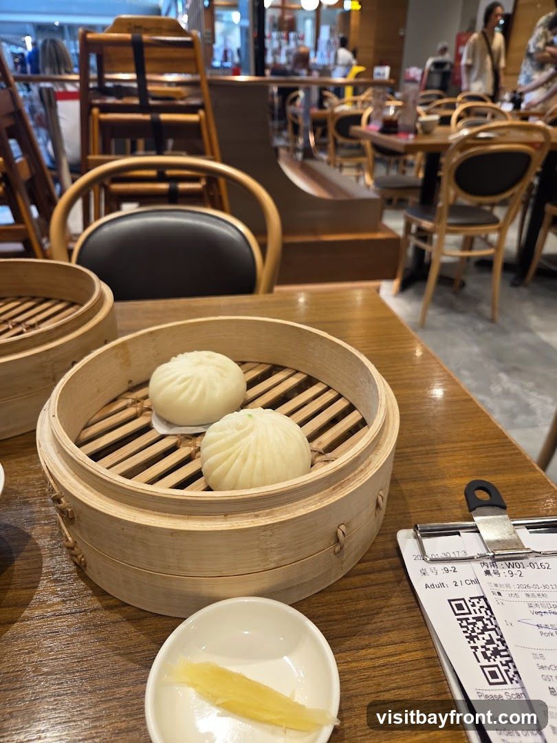 Two steamed buns in a bamboo steamer basket sit on a wooden table, next to a small dish of pickled ginger. A receipt is clipped to a clipboard in the foreground. The background shows a restaurant interior with wooden chairs and tables.
