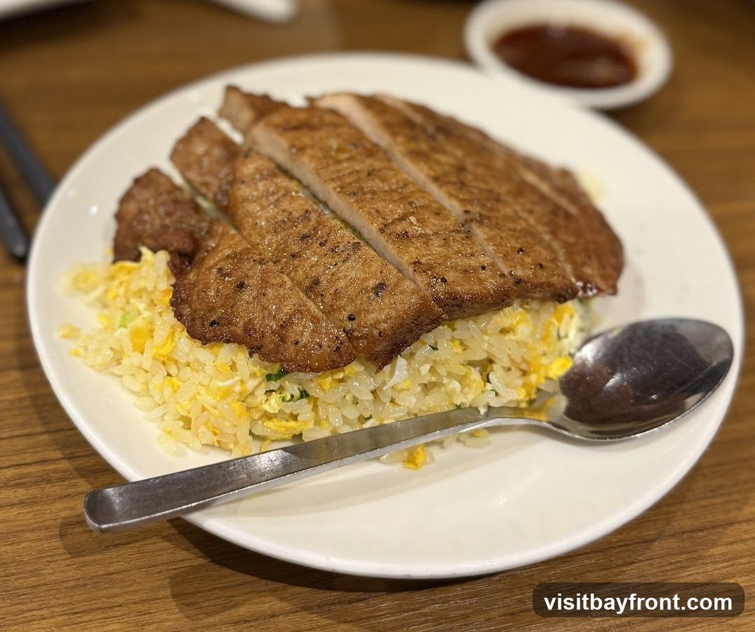 A plate of fried rice topped with sliced grilled pork chops. A spoon rests on the side of the plate. A small dish of sauce is visible in the background.