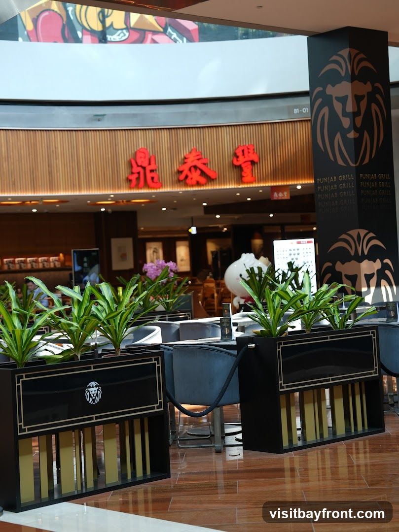 The image shows the interior of a restaurant with a prominent lion logo on a pillar. The pillar is black with a copper-colored lion face and repeated text "PUNJAB GRILL". In the background, red Chinese characters are visible above a wooden-paneled wall. The foreground features black planters with green plants, and tables with chairs are set up for dining. A large white cartoonish figure, possibly a mascot, is visible in the background.