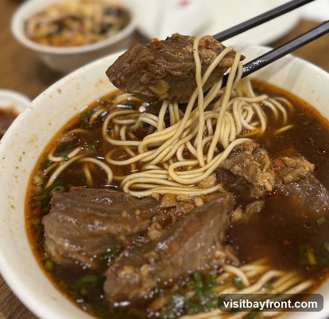 A close-up shot of a bowl of beef noodle soup. Noodles and chunks of beef are being lifted out of the broth with chopsticks. The soup appears to be rich and dark, garnished with chopped green onions. In the background, out of focus, are other bowls of food.