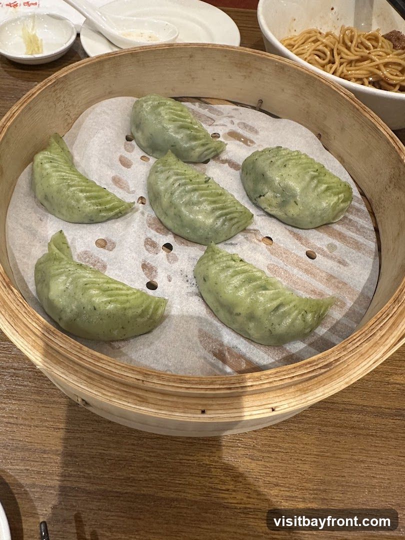 A bamboo steamer basket filled with six green dumplings, each with a pleated edge. The dumplings are arranged on white parchment paper. In the background, a bowl of noodles and small dishes are visible on a wooden table.
