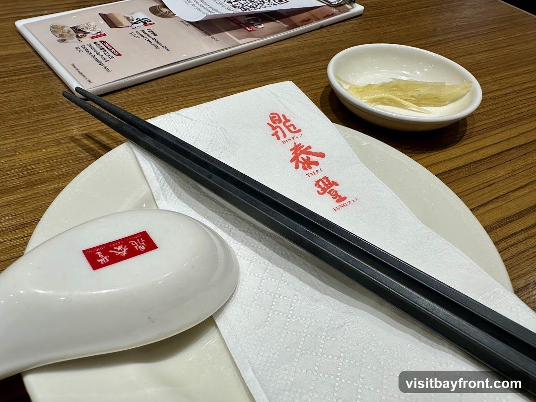 A table setting with a white plate, black chopsticks, a white spoon with red branding, a white napkin with red text, and a small dish of pickled ginger. A menu is partially visible in the background.