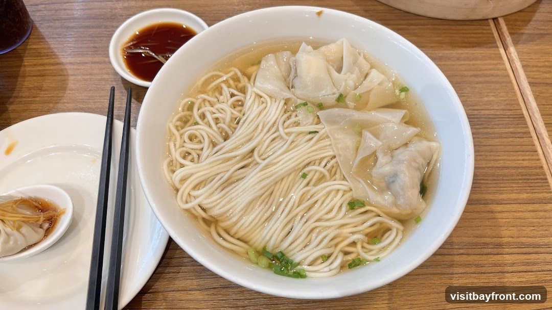 A bowl of wonton noodle soup with thin noodles and several wontons in a clear broth, garnished with chopped green onions. A small dish of dipping sauce and a spoon with a dumpling are visible on the side, along with chopsticks.