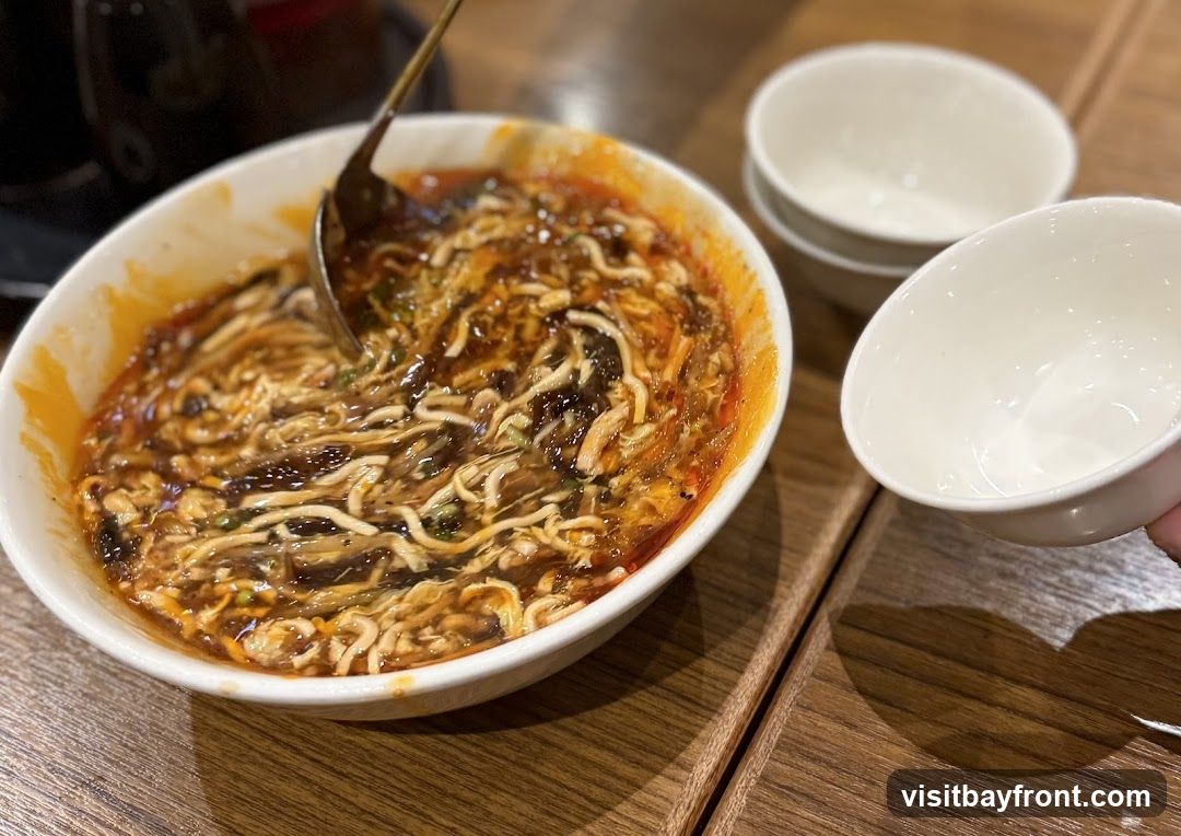 A close-up shot of a bowl of hot and sour soup, with noodles, vegetables, and a rich broth. A ladle is in the bowl, and two small white bowls are to the right. The soup appears to be served on a wooden table.