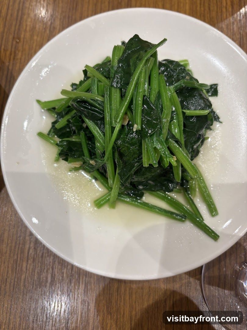 A close-up shot of sautéed spinach served on a white plate. The spinach is bright green and appears to be cooked with garlic and a light sauce. The plate is on a wooden table.