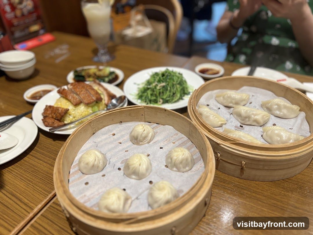 A table laden with various dishes, including two bamboo steamers filled with dumplings, a plate of fried rice topped with sliced meat, a dish of green vegetables, and small bowls of dipping sauce. A person is visible in the background, looking at their phone.