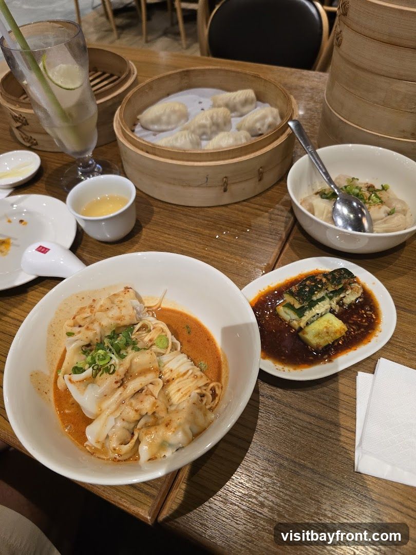 A table laden with various dishes, including steamed dumplings in bamboo steamers, a bowl of wontons in chili oil, a small dish of sliced cucumbers in sauce, and a refreshing drink with lime and lemongrass. The setting appears to be a restaurant with wooden tables and chairs visible in the background.