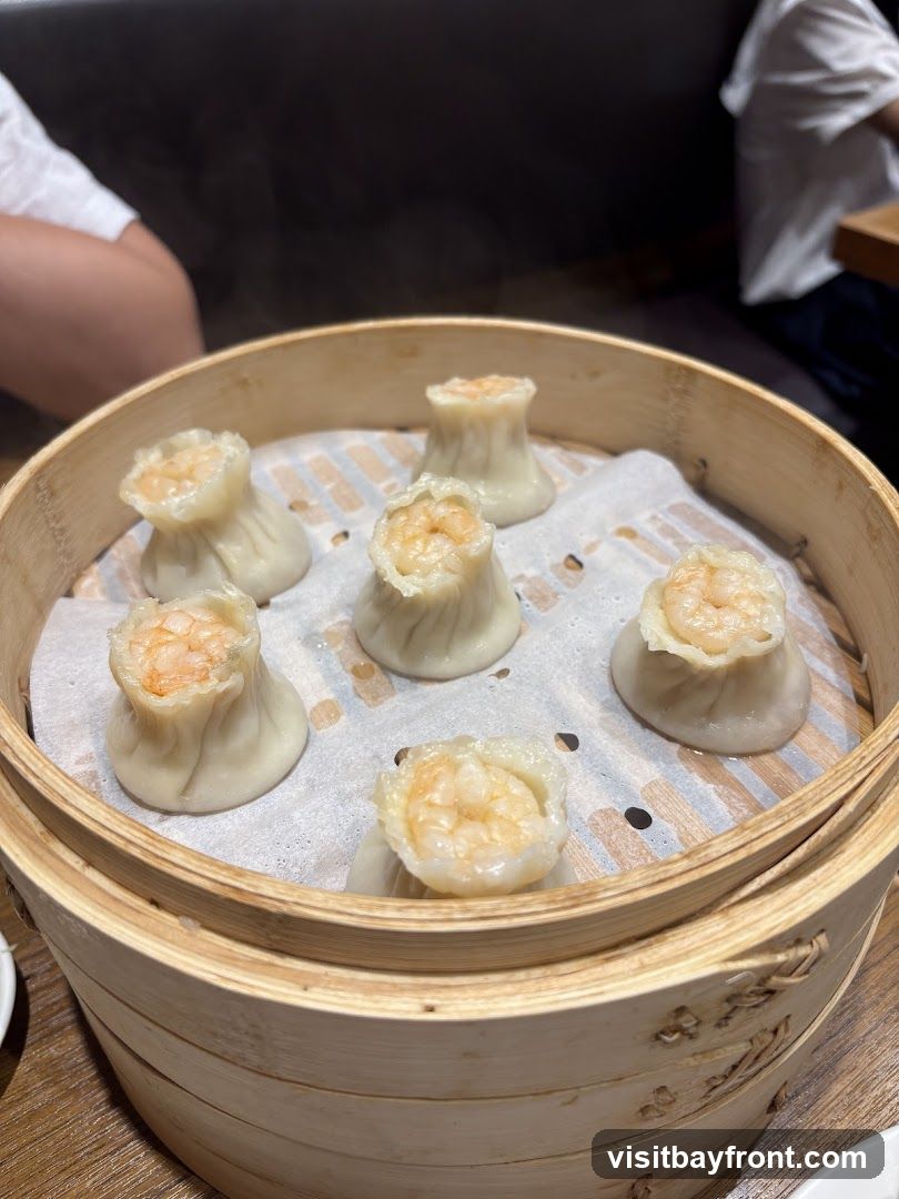 A bamboo steamer basket filled with six steamed shrimp dumplings, also known as siu mai. The dumplings have a delicate wrapper and are topped with a piece of shrimp. Steam is rising from the dumplings, indicating they are freshly cooked. The basket is placed on a table, and parts of diners are visible in the background.