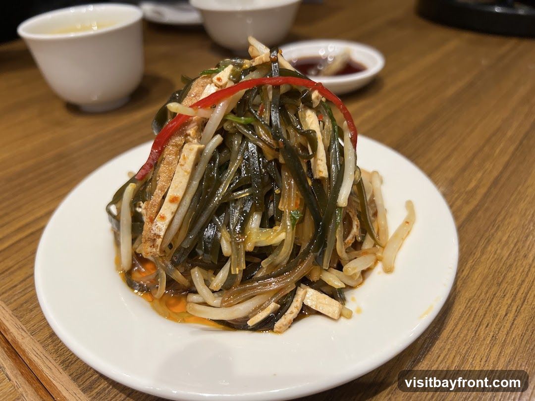 A close-up shot of a white plate filled with a refreshing seaweed salad. The salad is a mix of dark green seaweed strands, thinly sliced white tofu, and slivers of red chili pepper, all tossed in a light, savory dressing. In the background, two white ceramic bowls and a small dish of dark sauce are visible on a wooden table.