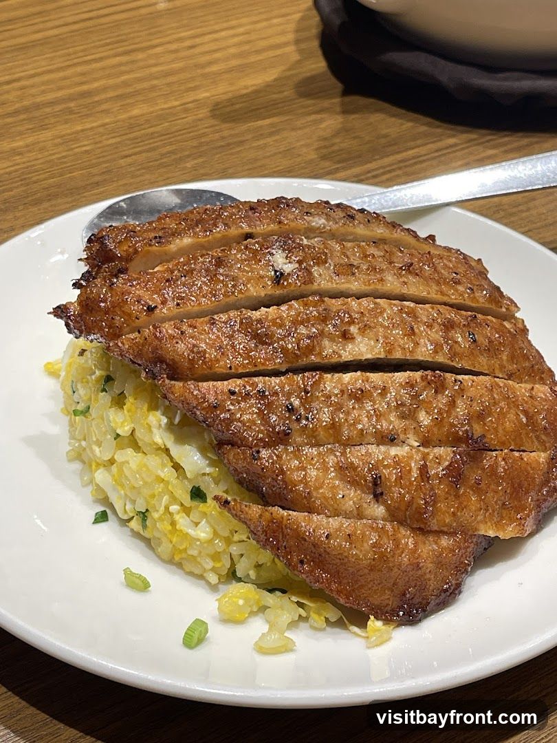 A close-up shot of a plate of fried rice topped with sliced grilled chicken. The fried rice appears to be made with scrambled eggs and chopped green onions. The chicken is golden brown and has grill marks. A spoon rests on the side of the plate.