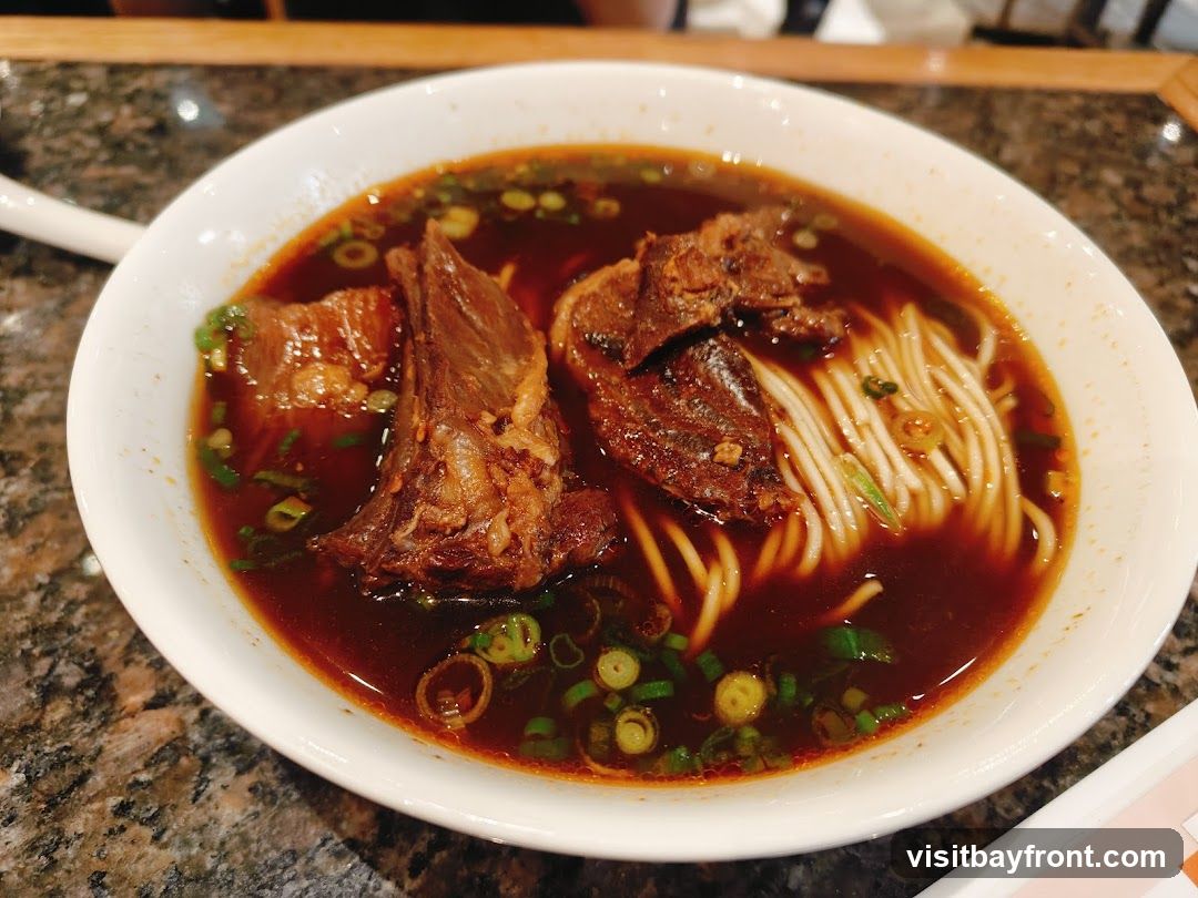 A close-up shot of a bowl of beef noodle soup. The soup is a rich, dark broth with large pieces of tender beef and thin noodles. Chopped green onions are sprinkled on top. The bowl is white and sits on a speckled countertop.