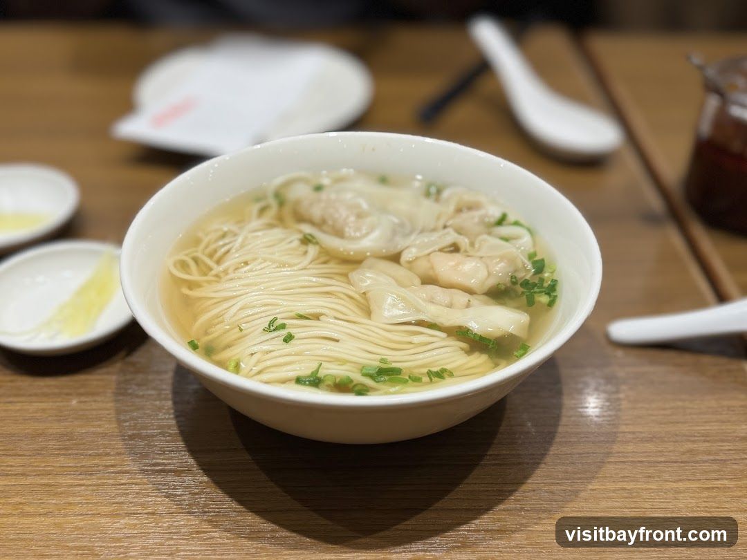 A close-up shot of a bowl of wonton noodle soup, garnished with chopped green onions. The soup is clear and light-colored, with thin noodles and several wontons visible. Two small white dishes containing what appears to be pickled vegetables or condiments are placed to the left of the bowl. In the background, out of focus, are white plates, spoons, and a jar of chili oil. The meal is served on a wooden table.