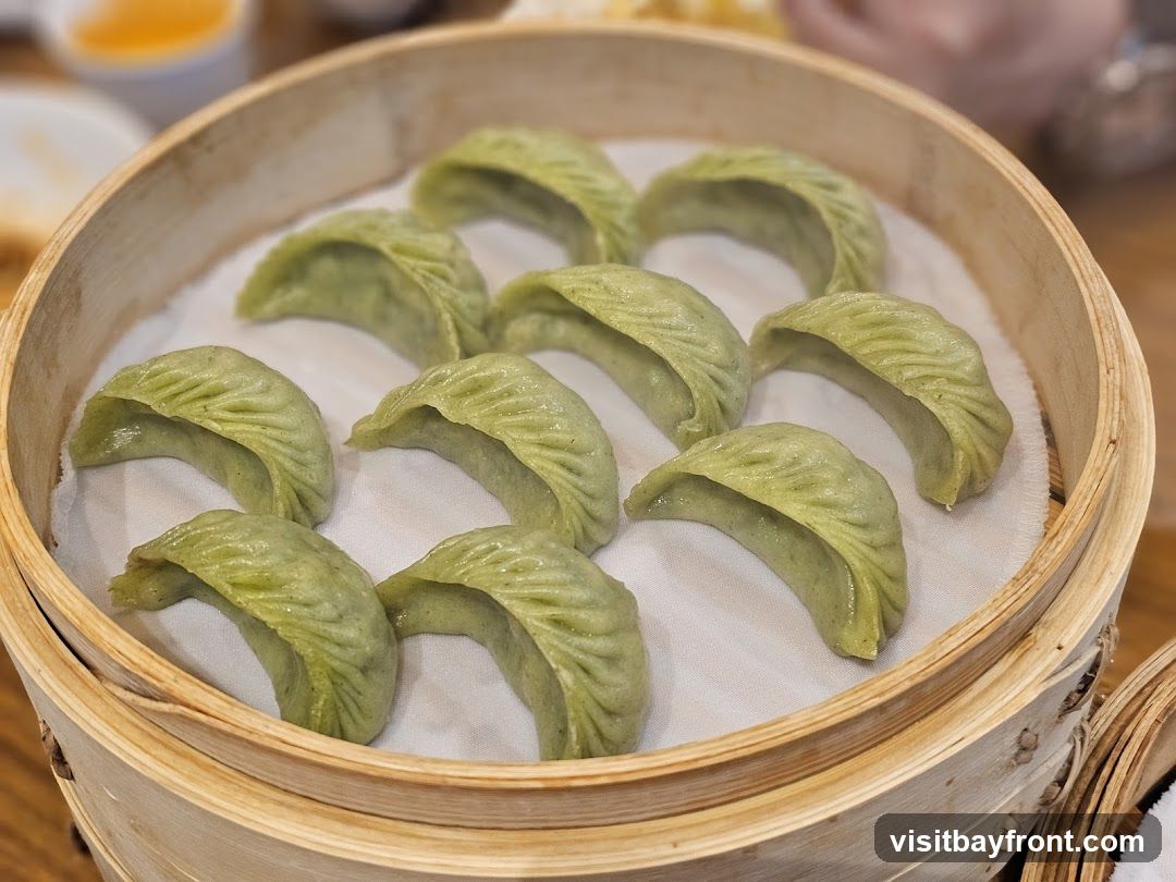 A close-up shot of several green dumplings arranged in a bamboo steamer basket. The dumplings have a pleated appearance and are resting on white parchment paper. In the background, out of focus, are hints of other dishes and tableware.
