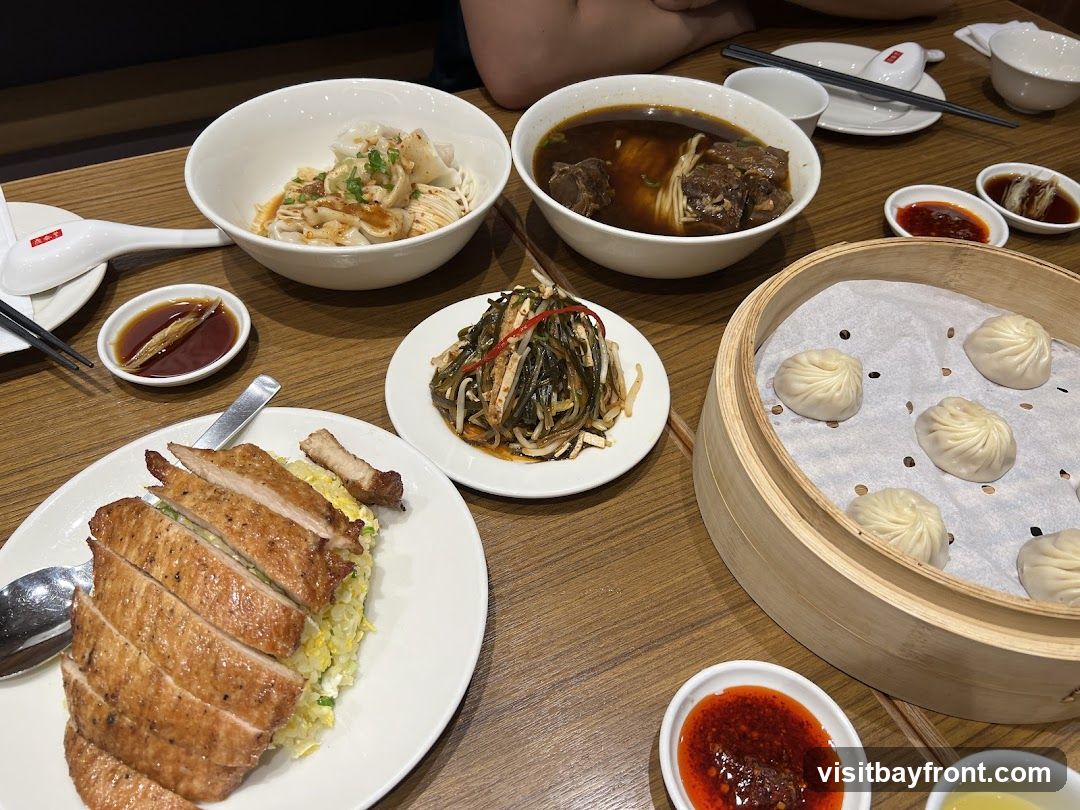 A table laden with various Chinese dishes, including a bowl of wonton noodles, a steaming bowl of beef noodle soup, a plate of sliced roasted meat over fried rice, a bamboo steamer basket filled with soup dumplings, and a side dish of pickled vegetables. Small dipping bowls of sauces are also visible.