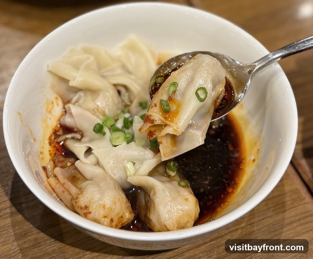 A close-up shot of a bowl of wontons in a spicy chili oil sauce, garnished with chopped green onions. A spoon is lifting a wonton from the bowl, showing the glistening sauce.