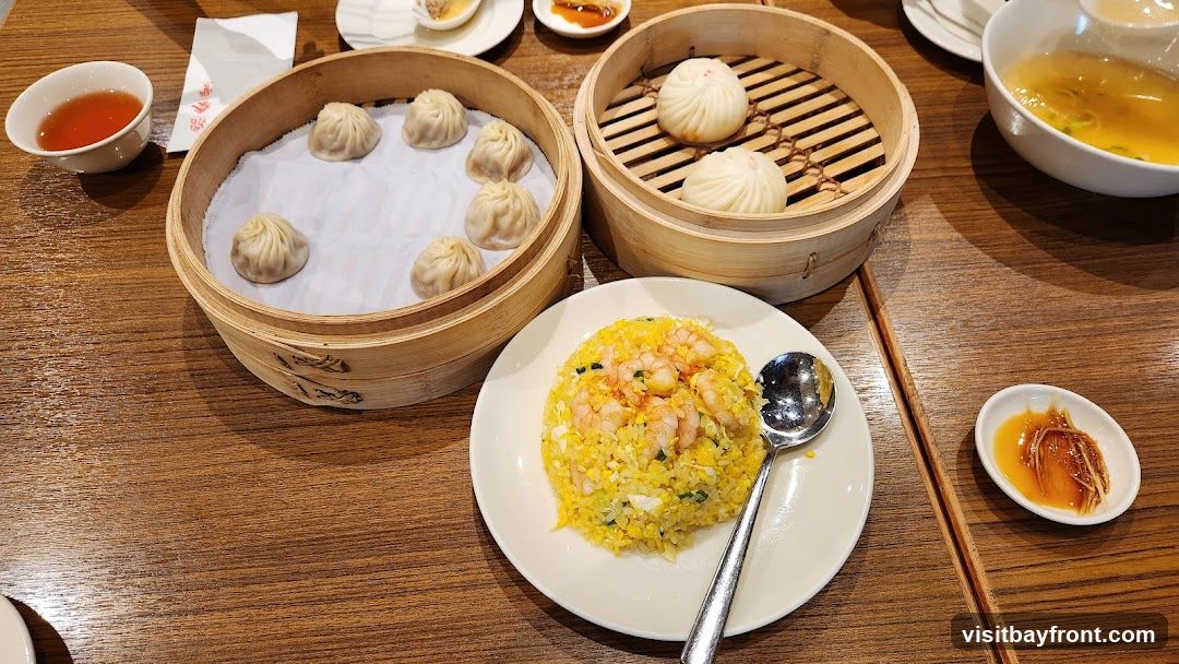 A table laden with a dim sum meal, featuring two bamboo steamers filled with dumplings, a plate of shrimp fried rice, a bowl of soup, and small dishes of dipping sauce and tea.