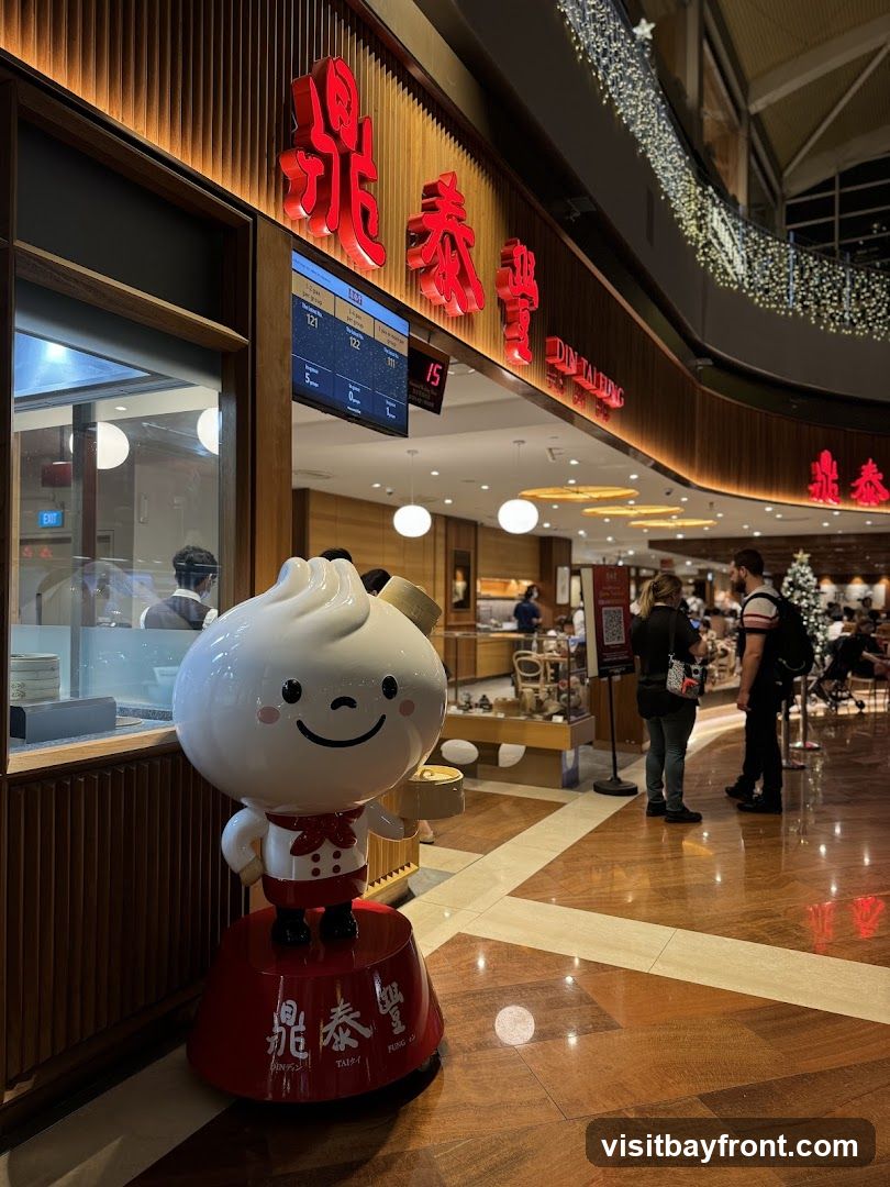 A cheerful dumpling mascot stands in front of a Din Tai Fung restaurant. The restaurant's red signage is visible above the entrance, along with a digital display showing queue numbers. People are seen entering and dining inside. The interior features warm lighting and wooden accents.