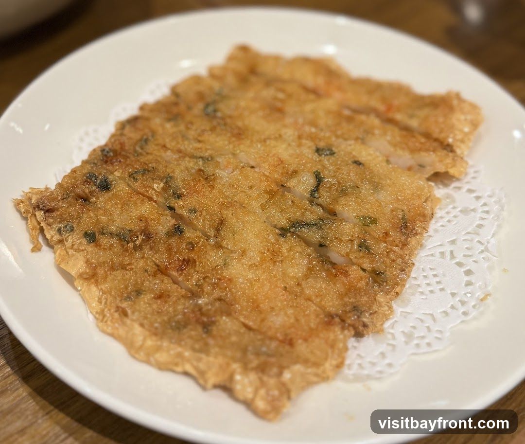 A close-up shot of a fried fish cake on a white plate. The fish cake is golden brown and appears to be made with minced fish and herbs. It is cut into rectangular pieces and served on a white doily.