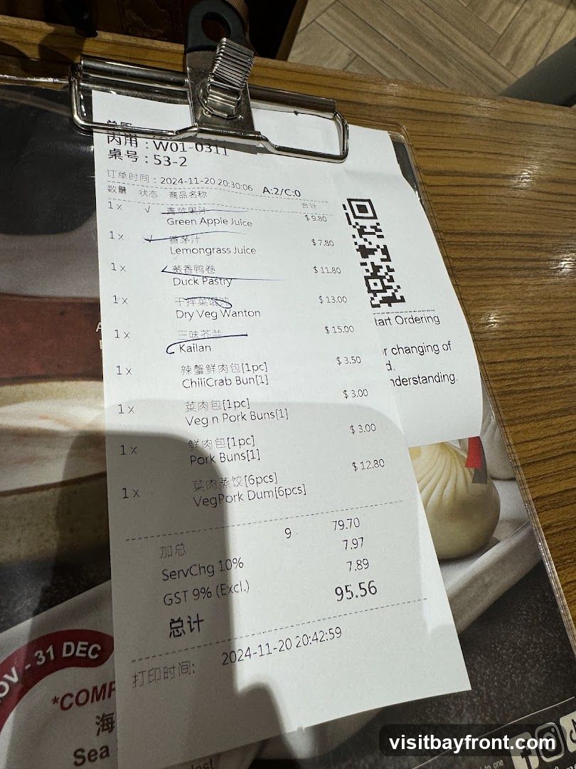 A close-up view of a receipt or order slip from a restaurant, detailing food items and their prices. The receipt is clipped to a clipboard and rests on a wooden table. Visible items include Green Apple Juice, Lemongrass Juice, Duck Pastry, Dry Veg Wanton, Kailan, ChiliCrab Bun, Veg n Pork Buns, Pork Buns, and VegPork Dum. The total amount is 95.56. A QR code for ordering is also visible on the right side of the receipt.