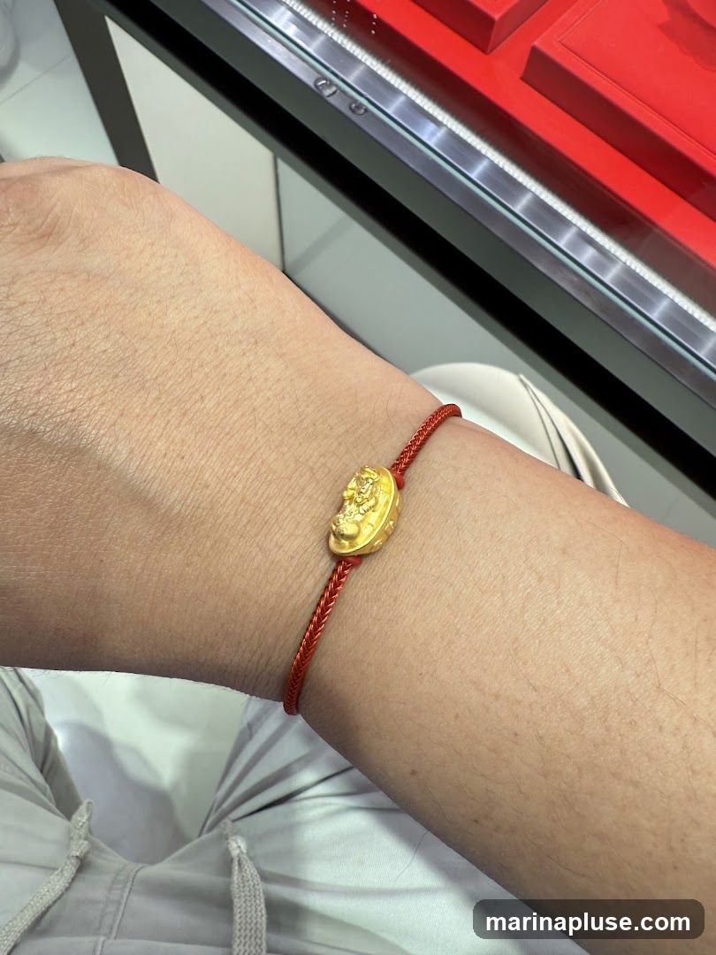 A close-up shot of a person's wrist wearing a thin red string bracelet with a small, oval-shaped gold charm. The charm appears to have an intricate design, possibly a deity or mythical creature, embossed on its surface. The background shows a blurred display case with red velvet lining, suggesting a jewelry store setting.