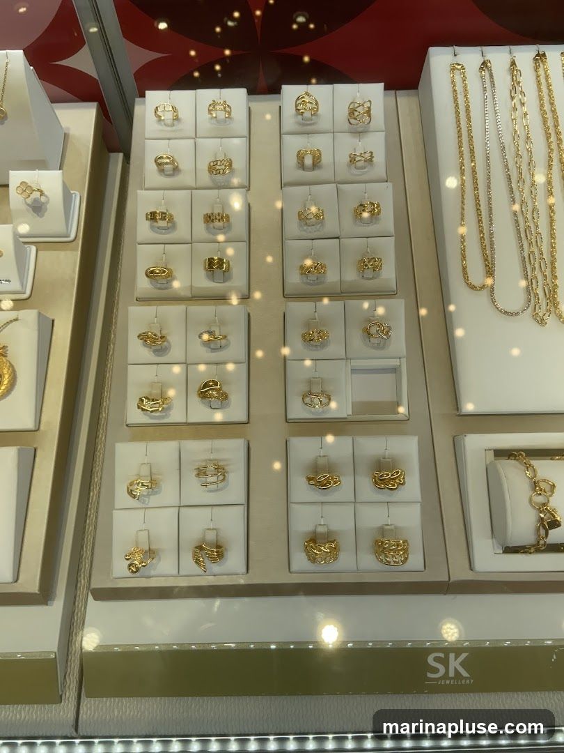 A display case filled with various gold rings and necklaces. The rings are arranged in grids on white stands, showcasing different designs. On the right, several gold chains are displayed vertically. At the bottom of the display, a logo reads 'SK JEWELLERY'.