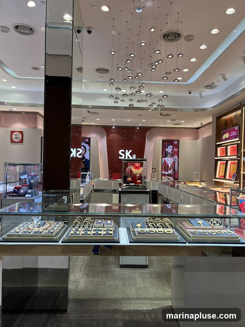The interior of a well-lit jewelry store. Display cases in the foreground showcase various gold necklaces, bracelets, and rings on black and red velvet stands. In the background, larger display cases hold more jewelry, and prominent signage with the letters "SK" is visible on a red wall. Decorative lighting fixtures hang from the ceiling.