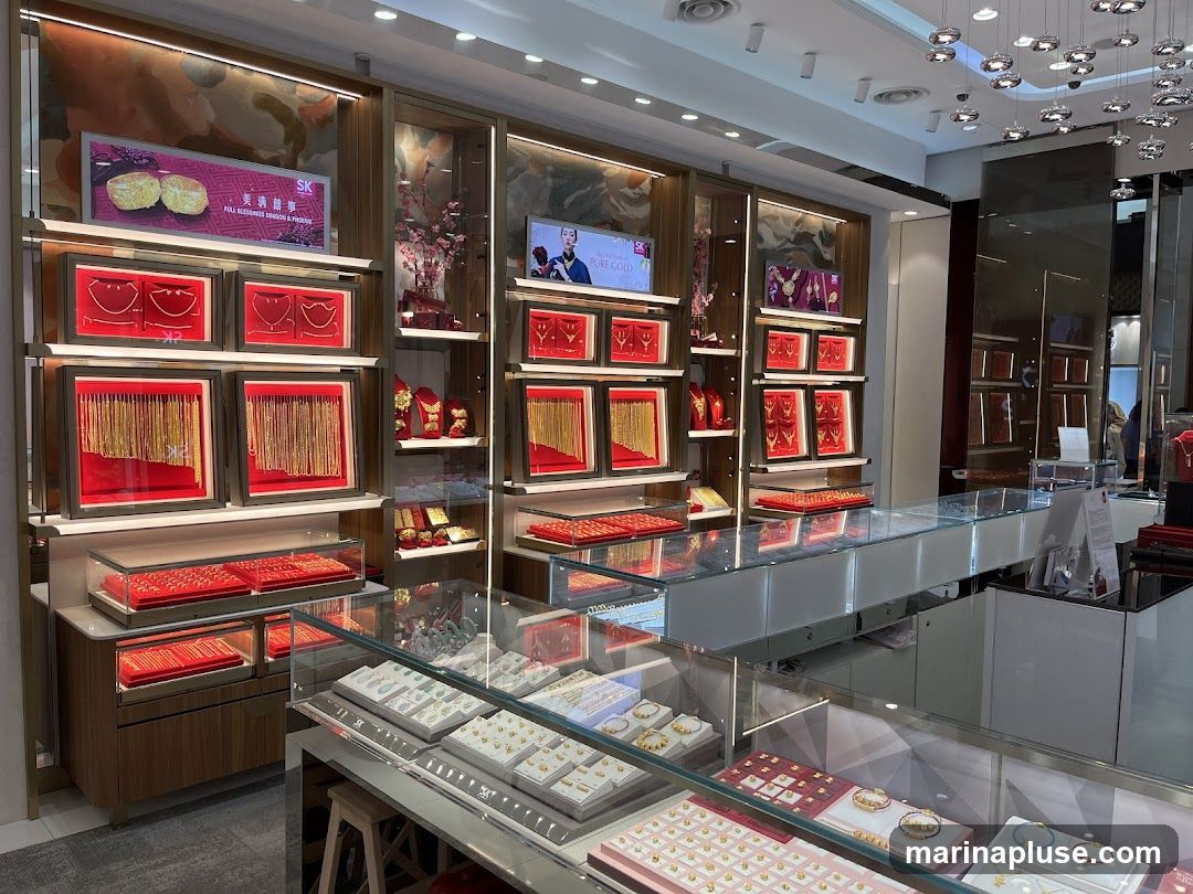 The interior of a jewelry store displays various gold necklaces, bracelets, and rings in illuminated display cases. The walls are adorned with framed jewelry and digital screens showing advertisements. The store features modern lighting fixtures and a clean, organized layout.