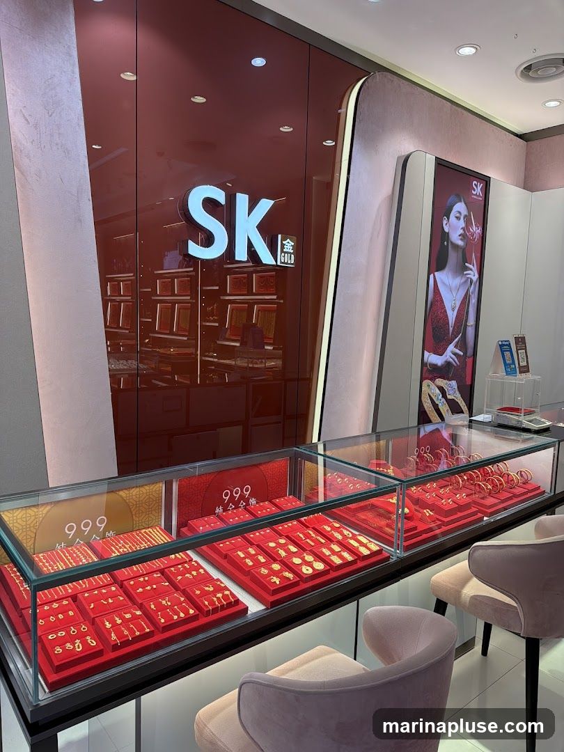 The interior of a jewelry store with a prominent "SK GOLD" sign. Display cases filled with gold jewelry, including necklaces, bracelets, and earrings, are arranged in front of a red, reflective wall. A digital advertisement featuring a model wearing jewelry is displayed on the right. Two plush chairs are placed in the foreground.