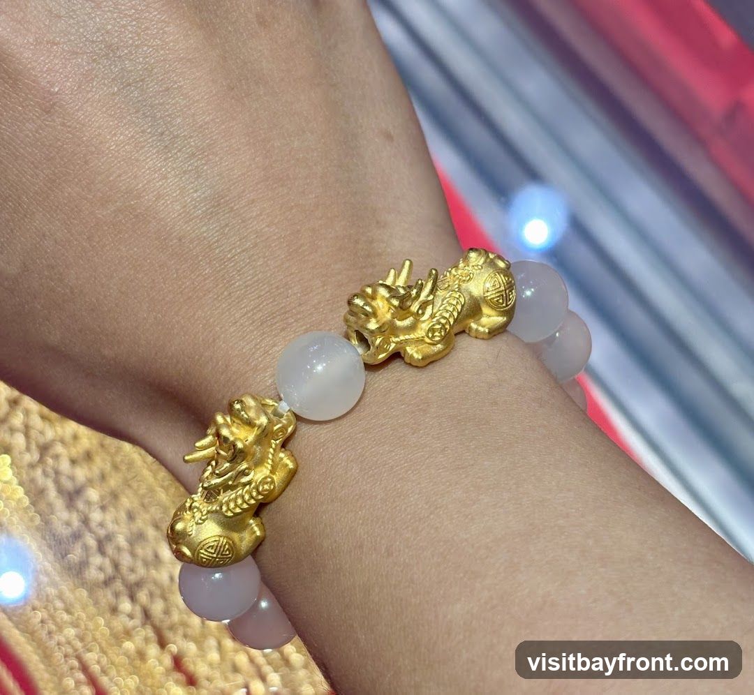 A close-up shot of a bracelet on a person's wrist. The bracelet features several gold charms shaped like mythical creatures, interspersed with smooth, light-colored beads. The background is blurred, showing hints of gold chains and a red and white striped pattern.