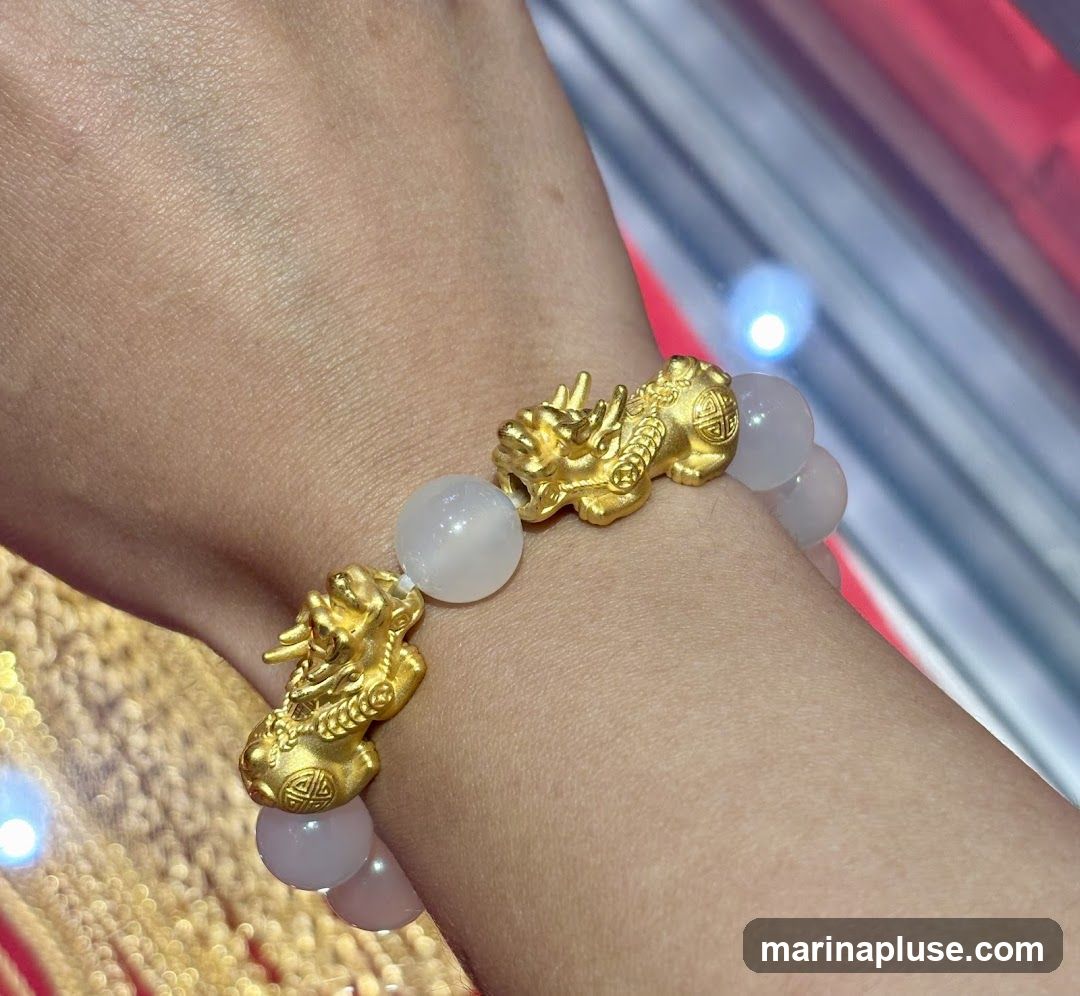 A close-up shot of a bracelet on a person's wrist. The bracelet features several gold charms shaped like mythical creatures, interspersed with smooth, light-colored beads. The background is blurred, showing hints of gold chains and a red and white striped pattern.