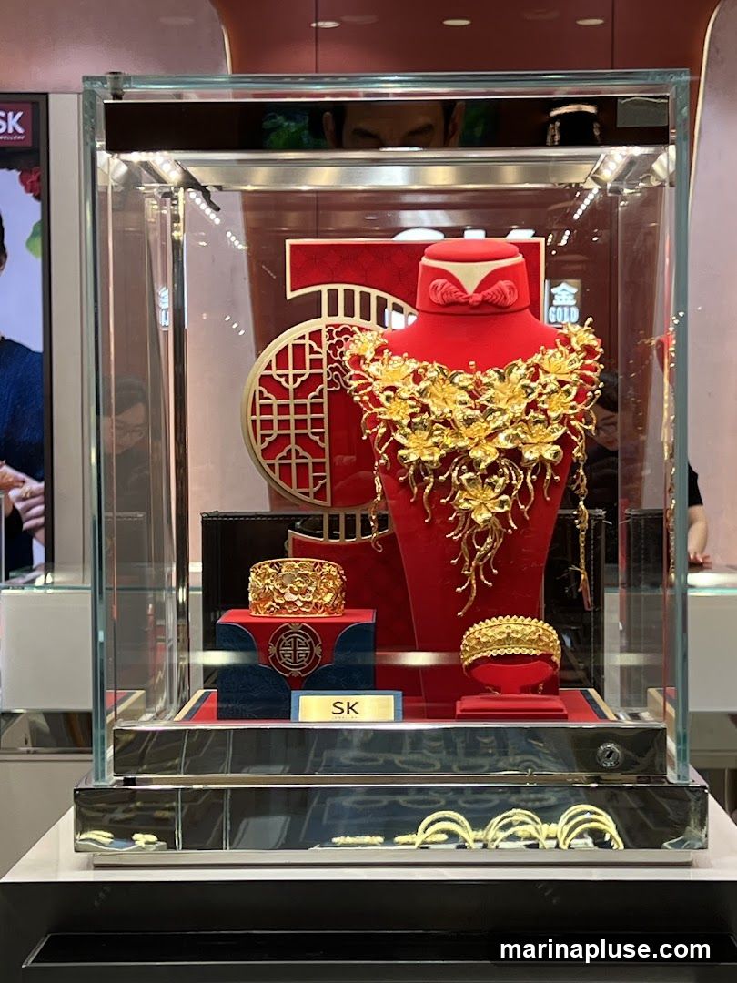 A display case filled with ornate gold jewelry, including a large floral necklace, two bracelets, and several rings. The jewelry is showcased on red velvet stands and platforms within a glass case. A red backdrop with a gold circular pattern and the number '50' is visible behind the necklace. The brand name 'SK' is displayed on a small plaque.