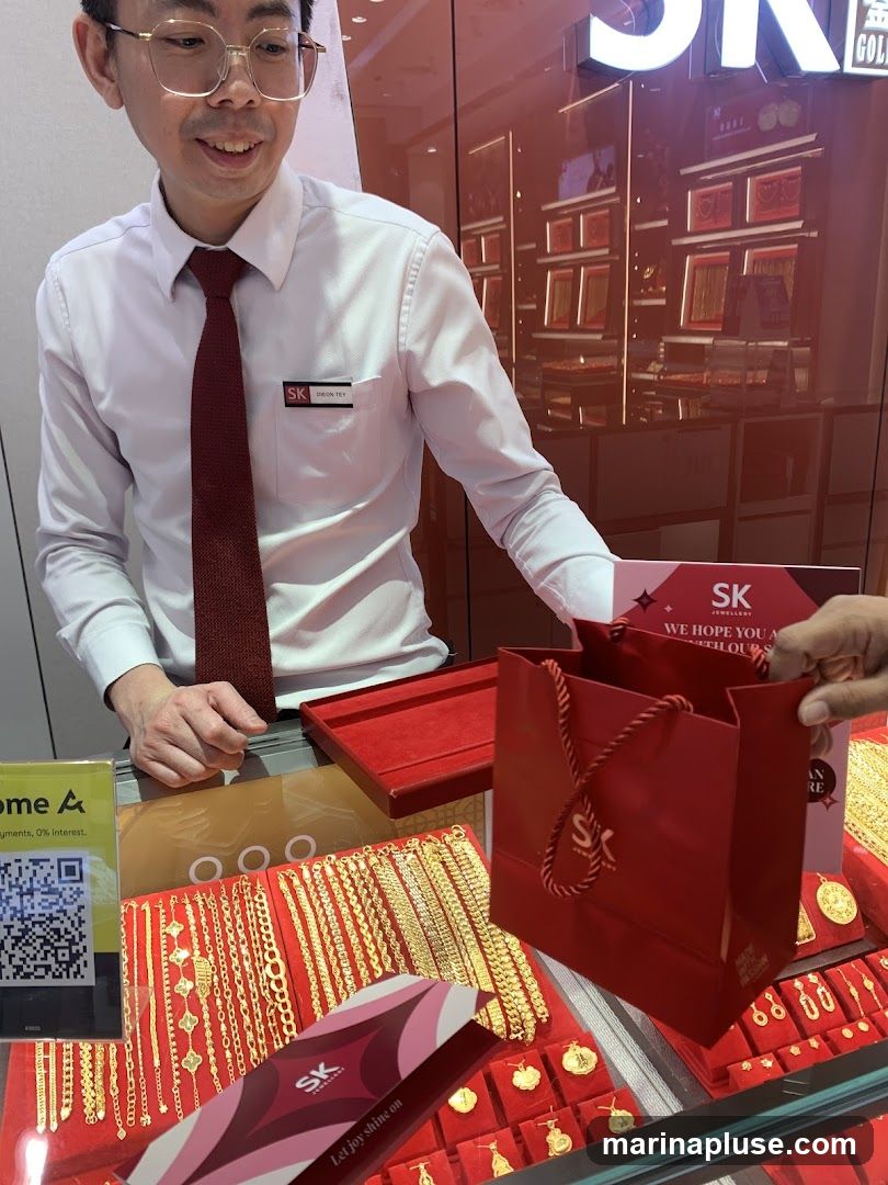 A store employee in a white shirt and red tie stands behind a counter displaying various gold necklaces and bracelets. A red shopping bag with the SK logo is being handed to a customer. The store's interior, with more jewelry displays, is visible in the background.