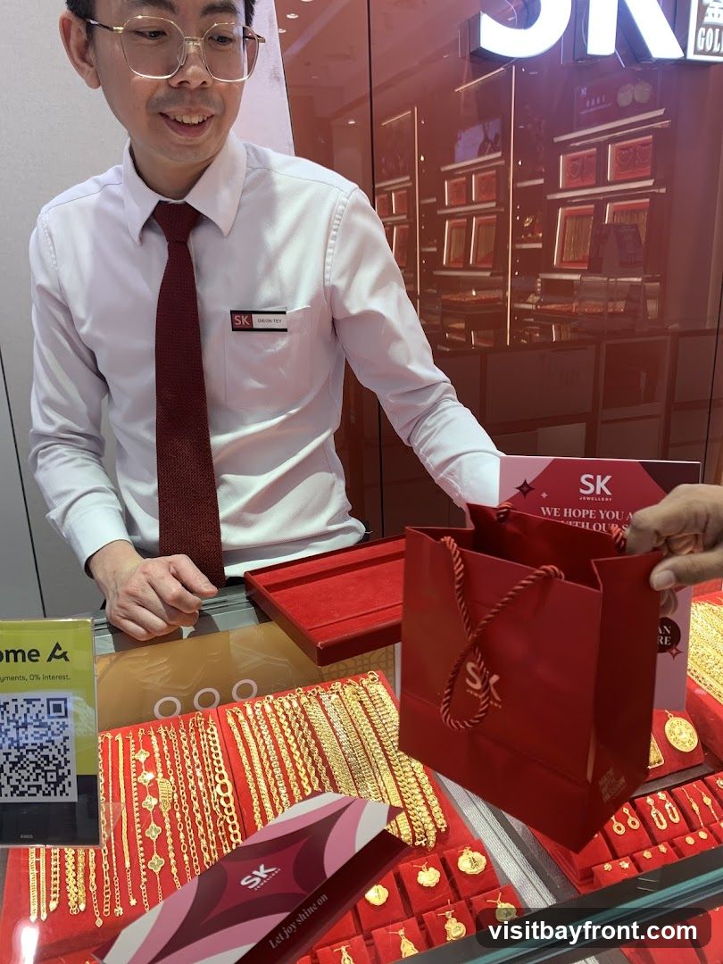 A store employee in a white shirt and red tie stands behind a counter displaying various gold necklaces and bracelets. A red shopping bag with the SK logo is being handed to a customer. The store's interior, with more jewelry displays, is visible in the background.