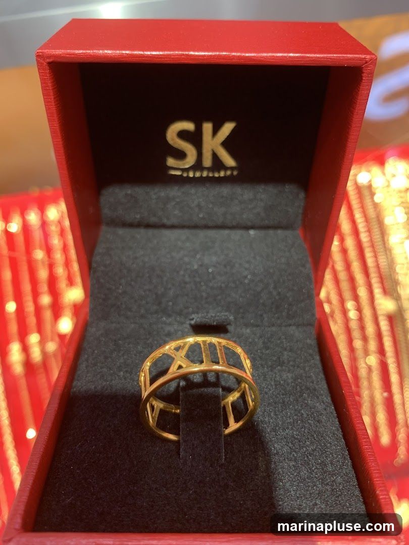 A gold ring with Roman numerals is displayed in a red jewelry box. The box has the logo "SK JEWELLERY" printed in gold on the inside lid. The background shows blurred images of gold necklaces or bracelets.