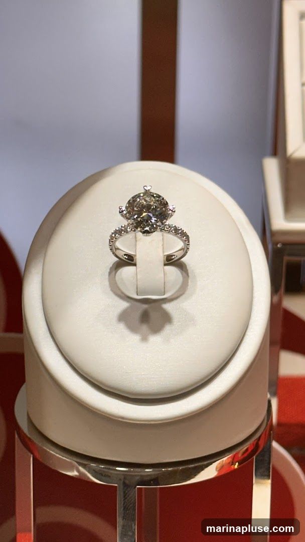 A large, oval-cut diamond ring with smaller diamonds on the band is displayed on a white, cushioned stand. The stand is on a metallic base. The background is blurred with vertical brown and white stripes and hints of red.
