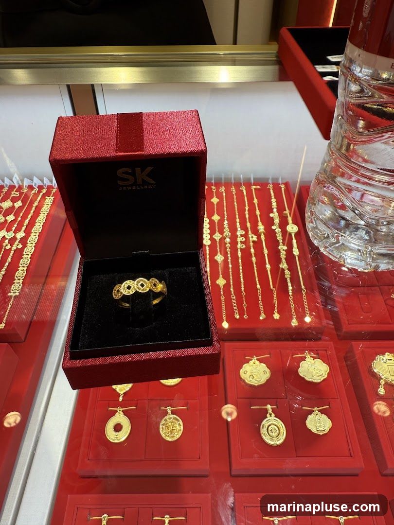 A close-up view of a jewelry display case featuring various gold items. In the foreground, an open red jewelry box displays a gold ring with a circular coin-like design. Behind the box, several gold bracelets with intricate patterns are arranged. Below, red display stands showcase gold pendants, some shaped like coins and others with decorative motifs. The overall presentation suggests a retail environment for fine jewelry.