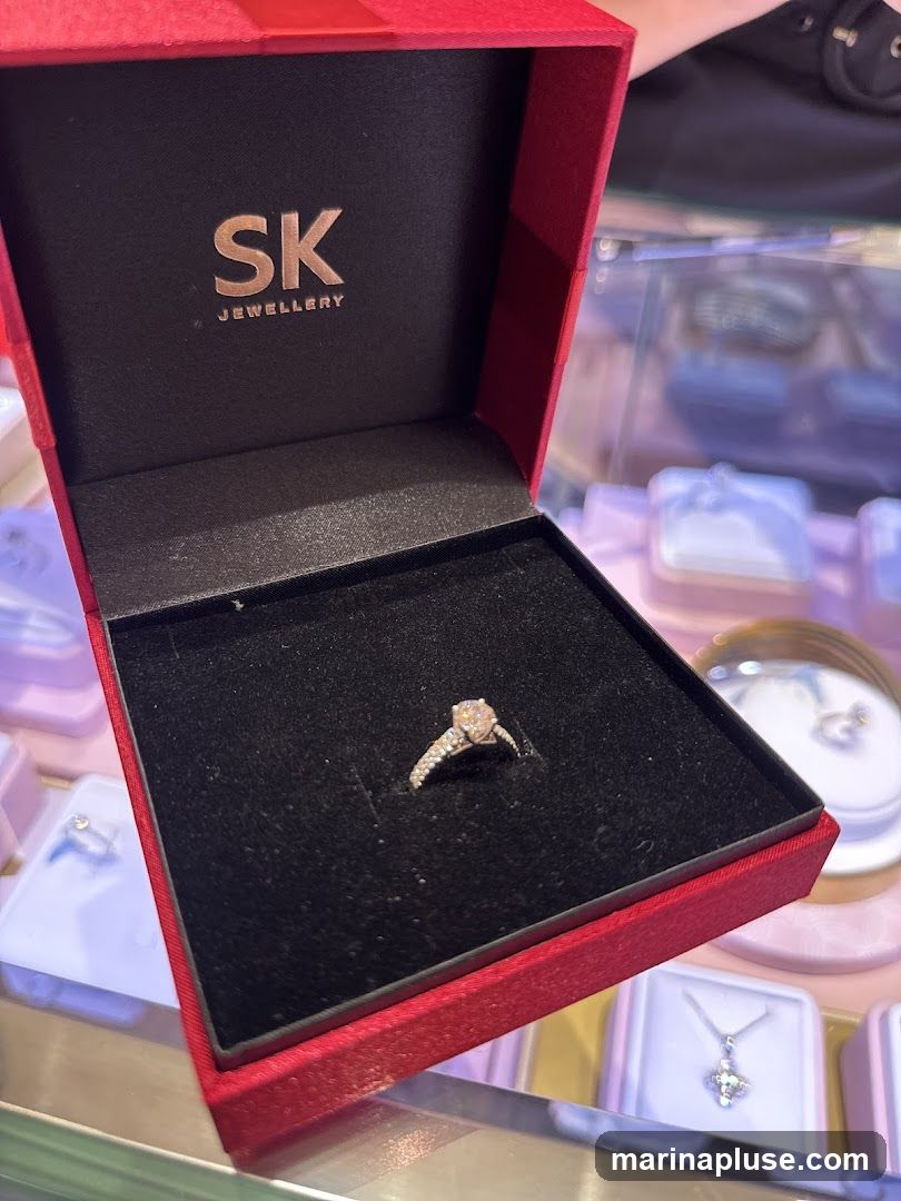 A close-up shot of a diamond engagement ring nestled in a red jewelry box. The inside of the box lid features the embossed logo "SK JEWELLERY". In the background, blurred display cases show other jewelry items, including necklaces and rings.
