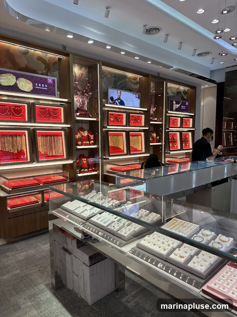 The interior of a well-lit jewelry store, showcasing a variety of gold necklaces, bracelets, and rings displayed in glass cases and on shelves. A staff member is visible behind the counter. Digital screens display advertisements for gold jewelry.