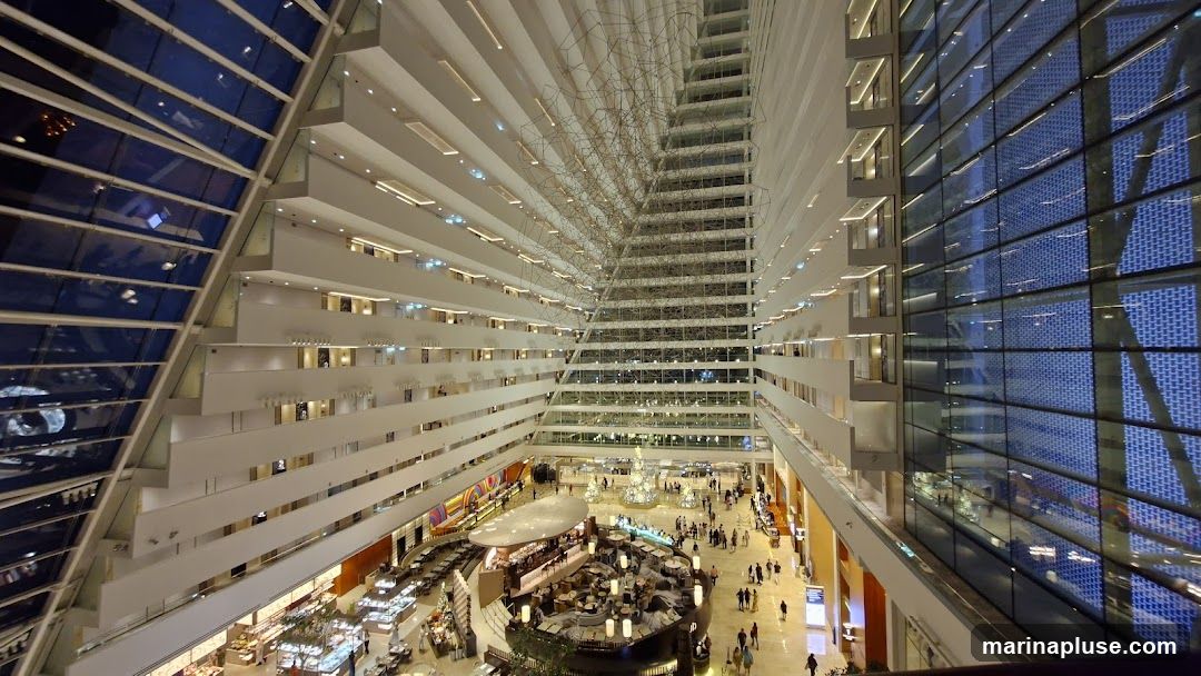 The image shows the interior of a vast atrium with many levels of balconies overlooking a central area. The architecture features a modern design with white walls and glass facades. In the center, there is a circular seating area with tables and chairs, surrounded by shops and walkways. People are seen walking around the atrium. A large, abstract sculpture hangs from the ceiling.