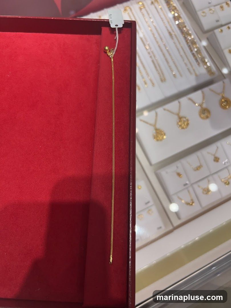 A close-up view of a delicate gold chain necklace displayed in a red velvet jewelry box. The necklace has a small spherical charm at the top and a fine, snake-like chain. In the background, a display case filled with various gold necklaces and pendants is visible. A tag attached to the necklace reads '916 3.17g'.