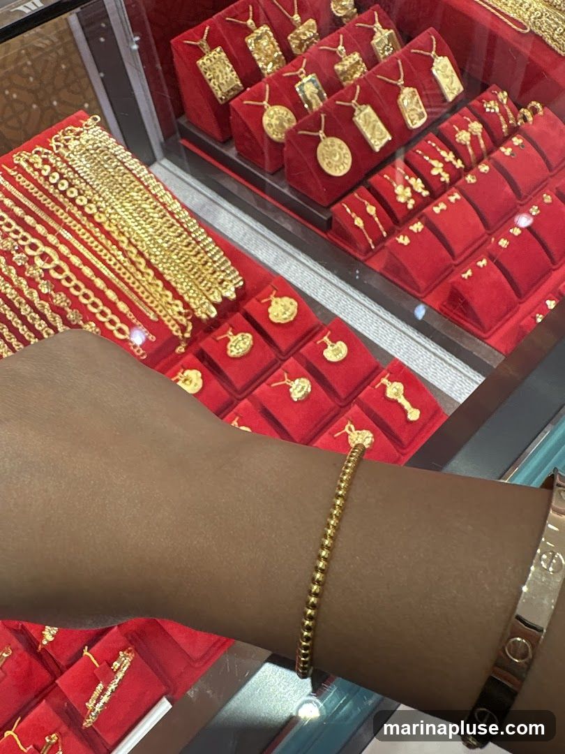 A close-up view of a jewelry display case filled with various gold necklaces, bracelets, and pendants. The items are arranged on red velvet stands. A person's arm is visible in the foreground, wearing a gold beaded bracelet and another gold bracelet with engraved logos.