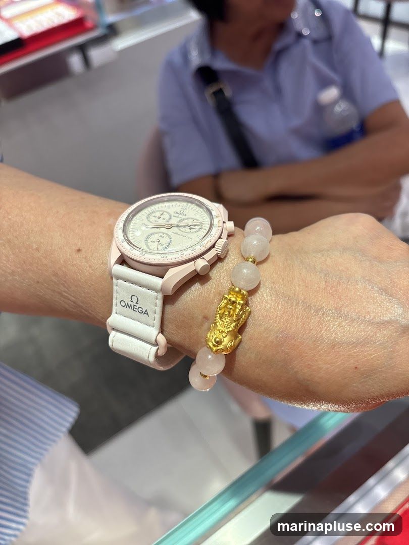 A close-up shot of a person's wrist wearing a light pink Omega watch and a bracelet with rose quartz beads and a gold charm. The watch has a white strap with the Omega logo. In the background, a person in a blue shirt is visible.