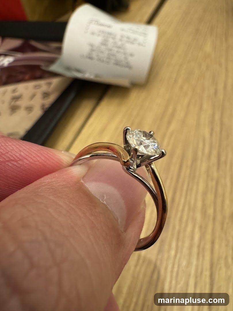 A close-up shot of a diamond engagement ring being held by a thumb and index finger. The ring features a large, round-cut diamond set in a two-tone metal band. In the background, a receipt or tag with text is visible.