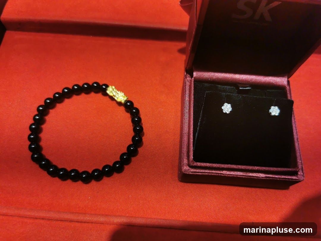 A black beaded bracelet with a gold dragon charm is displayed next to an open jewelry box containing a pair of diamond stud earrings. The box has the logo "SK" visible inside the lid. The items are presented on a red velvet surface.