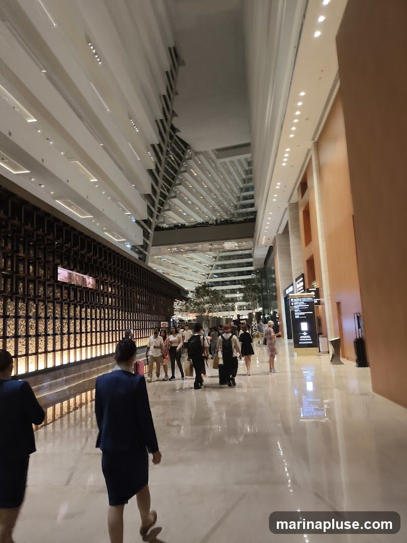 A wide, modern hotel lobby with a high ceiling and reflective floors. People are walking through the lobby, some appearing to be staff in uniform. The architecture features geometric patterns and warm wood tones.