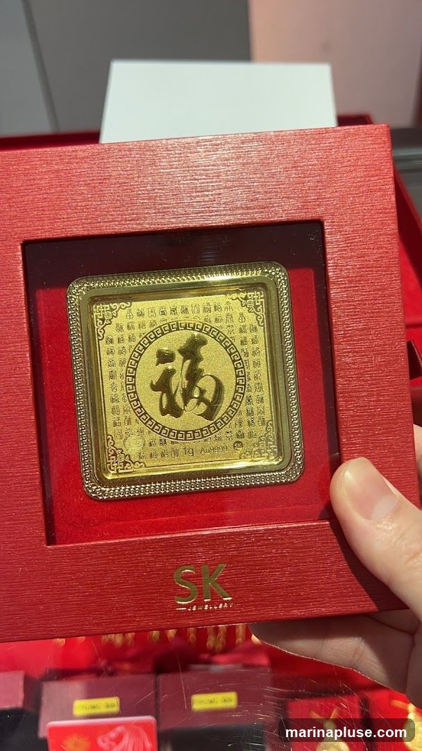 A close-up shot of a gold bar, encased in a red presentation box. The gold bar features intricate Chinese calligraphy, with a prominent character in the center, and smaller characters surrounding it. Text below the central character reads '1g Au9999'. The box has a textured red finish and a gold logo that appears to be 'SK JEWELRY'. The background shows other items in display cases, suggesting a retail environment.